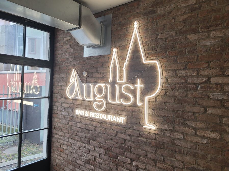 Der Schriftzug „August“ wurde als filigrane Neon-Optik Leuchtschrift gefertigt und strahlt gleichmäßig an der Wand. Besonders markant ist die integrierte Gestaltung: Eine feine Silhouette eines Weinglases sowie die Stadtkirche mit LED-Neon als Lichtkontur. sind harmonisch in den Logonamen eingebunden.