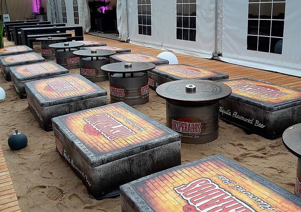 Lounge Möbel mit Strandfeeling mit Desperados-Öl-Fass-Tische und gepolstereten Sitzkisten laden Gäste zum verweilen ein.  Die Kombination aus rustikalem Industrie-Vintage Look des Fasses und warmem Holz schafft einen modernen, urbanen Stil mit sommerlichem Flair. Die Verbindung aus Lounge-Möbeln natürlichen Dekorationselementen und dem individuell gestalteten Fass-Stehtisch, unterstützt gezielt die Aufenthaltsqualität.