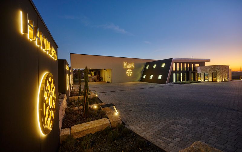 Lichtwerbung für das Weingut Manz aus Rheinhessen. Die drei Stelen mit durchgesteckten, leuchtenden vergoldeten-Metall-Profilschriften und dem golden hinterleuchteten Wappen präsentieren die Markenwerte „Handwerk, Leidenschaft, Faszination“ nachts mit maximaler Fernwirkung. Am Hauptgebäude des Weinguts ist der goldene „MANZ“-Schriftzug und Wappen an der hellen Fassade zu sehen.
