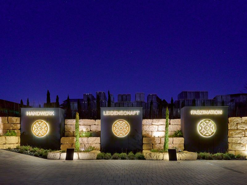 Drei schwarze Stelen vor einer Natursteinmauer leuchten nachts mit warmweißen Schriftzügen und goldenen Logos.Die Begriffe „Handwerk“, „Leidenschaft“ und „Faszination“ stehen zentral über dem Familienwappen.Eine moderne Lichtinstallation, die das Branding des Weinguts Manz nachts hervorhebt.