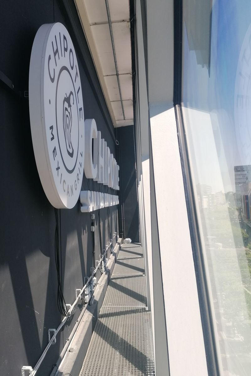 Die von uns installierte Werbeanlage der Marke Chipotle ist im Innenbereich der MyZeil. Die Besonderheit: Die Anlage ist so positioniert, dass sie durch die charakteristische Glasfassade des Centers nach außen strahlt. Das Bild verdeutlicht die professionelle Montage der Profilbuchstaben und des Rundlogos auf einer speziellen Unterkonstruktion.