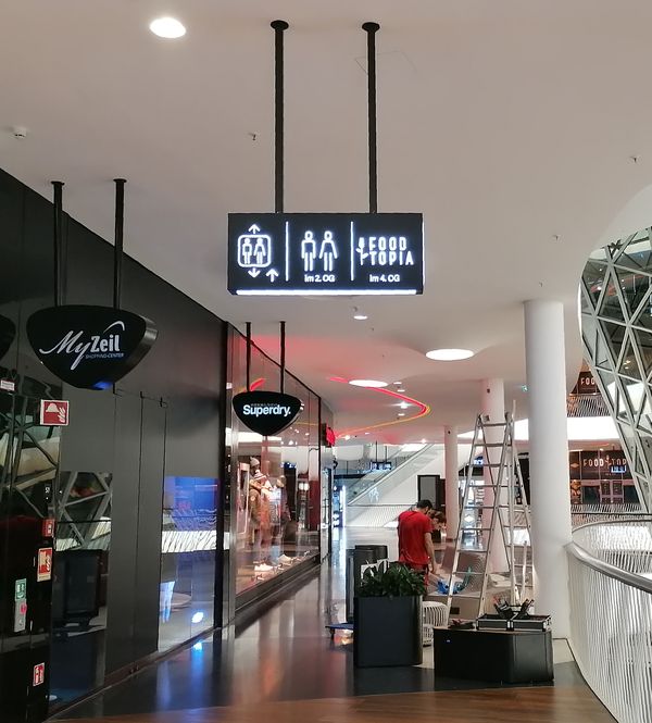 Wegeleitung mit leuchtende Piktogramm-Schildern im MyZeil. Der doppelseitige Ausleger präsentiert klar verständliche Piktogramme für Fahrstühle, WCs und den Food Court 