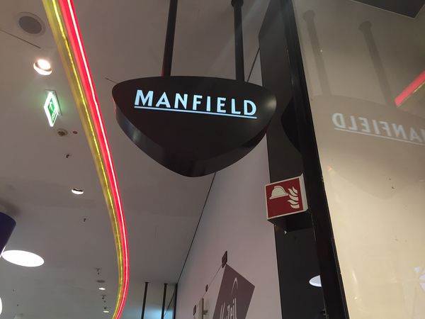 Die Shop-Beschilderung für Manfield zeigt, wie wichtig einheitliche Werbe-Ausleger für die Orientierung im Center sind. Dieser beidseitig leuchtende Ausleger in der center-spezifischen Sonderform garantiert, dass der Store bereits aus der Ferne im Laufgang optimal wahrgenommen wird. Die Lichttechnik ist auf Langlebigkeit und Energieeffizienz ausgelegt.