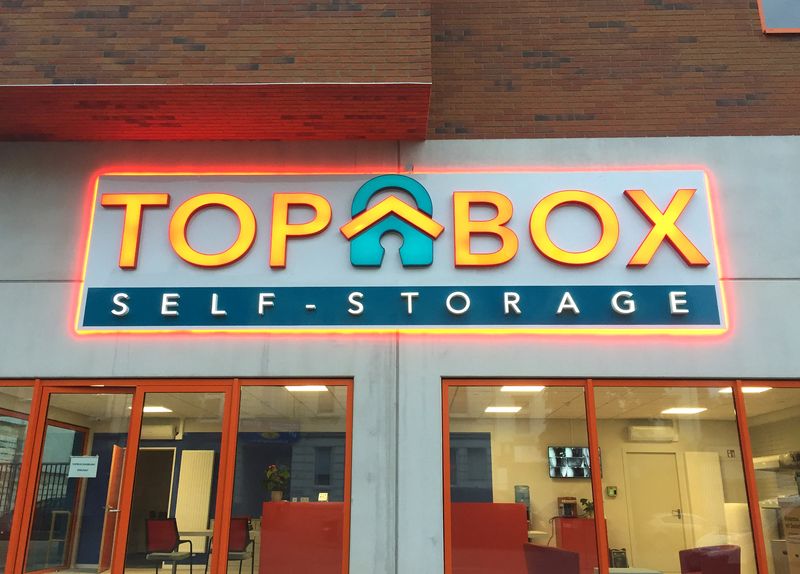 Eine Signalstarke Außenwerbung für Top-Box. Die Kombination aus Frontleuchtern und farbiger Konturbeleuchtung garantiert maximale Aufmerksamkeit im Stadtbild. Diese Lichtwerbeanlage nutzt kontrastreiche Farben (Orange und Blau) und eine zusätzliche umlaufende Neon-Kontur, um die Marke „Top Box