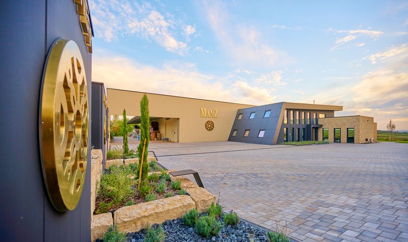 Aussenwerbung für das Weingut Manz aus Rheinhessen. Die drei Stelen mit vergoldeten-Metall-Profilschriften mit Rückbeleuchtung und Logo-Sign präsentieren die Markenwerte „Handwerk, Leidenschaft, Faszination“ am Vorplatz. Am Hauptgebäude des Weinguts ist der goldene Manz-Schriftzug und Logo an der sandfarbigen Fassade zu sehen.