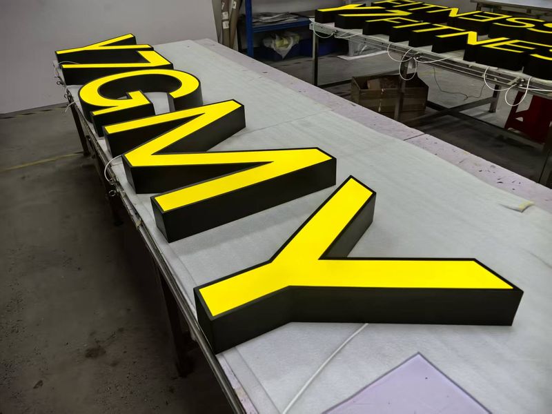 Großformatige Vollreliefbuchstaben für das Y7 GYM-Logo (gelbe Fronten, schwarze Zargen) liegen auf einem Produktionstisch zur finalen Montage oder Qualitätskontrolle. Die passgenau gefertigten Vollreliefbuchstaben bzw. Leuchtbuchstaben für das Fitnesstudio ist eine Kombination aus leuchtend gelben Fronten und edlen schwarzen Zargen, die für ein dreidimensionales, hochwertiges Erscheinungsbild mit markanter Seitenwirkung sorgt.